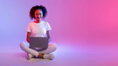 A smiling woman with curly hair uses a laptop while sitting with crossed legs against a gradient background clipart
