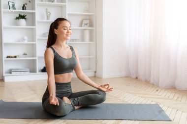 Huzurlu Asyalı kadın gözlerini kapatıp evde meditasyon yapıyor. Güzel, genç Koreli kadın, minimalist oturma odasının iç tarafındaki gri paspasın üzerinde oturuyor.
