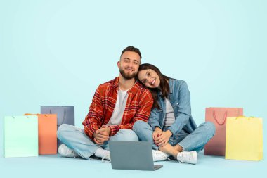 Dijital Ticari. Genç Avrupalı çift dizüstü bilgisayarlarıyla online alışveriş yapmayı, yerde alışveriş torbalarının arasında oturmayı, web sitelerine bakarken kameraya gülümsemeyi, mavi stüdyo arka planlarını seviyorlar.