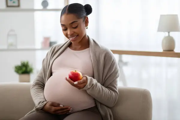 妊娠している間の栄養 ポジティブな健康期待の若いアフリカ系アメリカ人女性がリンゴを食べ 自宅で居心地の良いリビングルームでソファーに座って スナック コピースペースを持っています — ストック写真