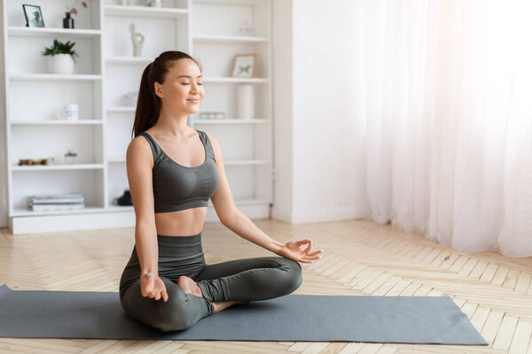 Serene asian woman closing her eyes and meditating at home, beautiful young korean female sitting in simple cross-legged position on gray mat in minimalist living room interior, copy space