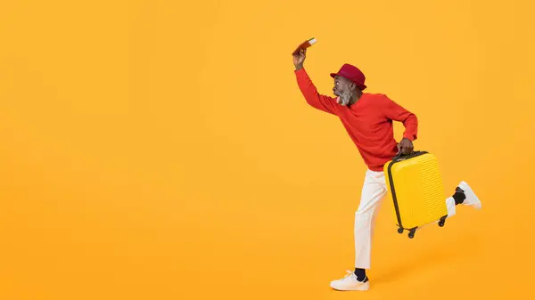 身穿红色毛衣 头戴黄色行李箱的精力充沛的资深黑人男子举着登机牌 带着在橙色背景下旅行的兴奋心情 工作室 — 图库照片
