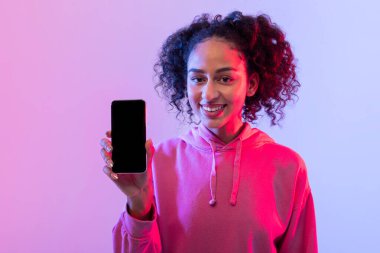 Pembe kapüşonlu genç bir kadın renkli arka planı olan kameraya akıllı telefon ekranı gösteriyor.