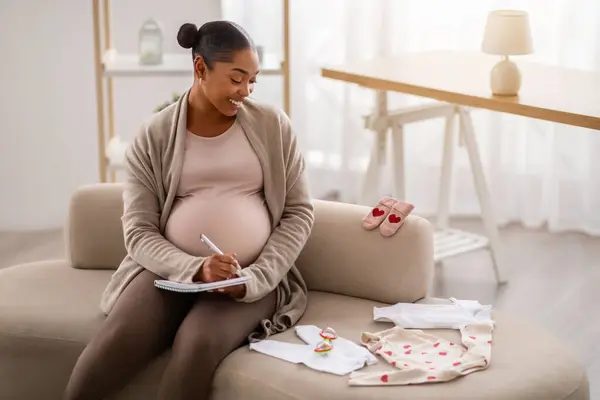 自宅でソファーに座って 赤ちゃんの服を見たり リストを作ったり 母性や労働の準備をしたり スペースをコピーしたりする幸せな若い黒人妊婦 — ストック写真