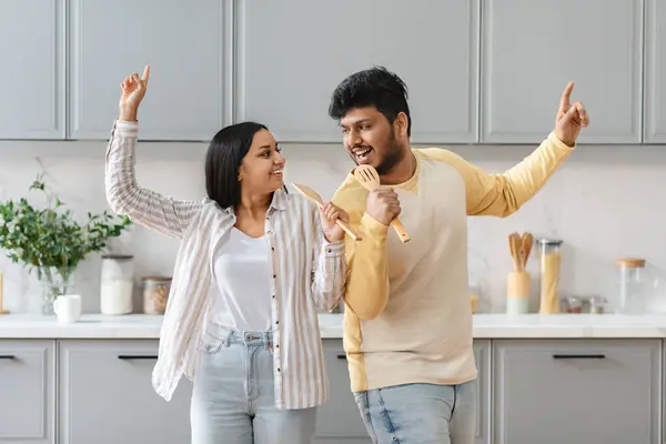 Funny young eastern couple singing and dancing together at kitchen, happy indian man and woman using utensil as microphone, performing while cooking at home