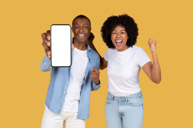 Muzaffer Afro-Amerikan çifti neşeli bir akıllı telefon ekranı gösterirken erkek, kadının sarı bir arka plan karşısında ürün sunumu için mükemmel tezahürat yaparken onu tutuyor.