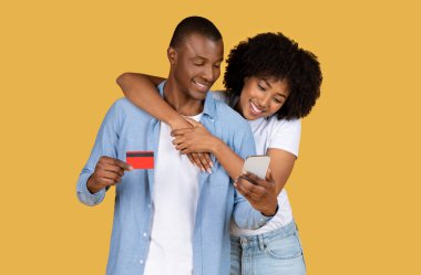Gülümseyen Afrikalı Afrikalı Amerikalı çift online alışveriş yapıyor, kredi kartı ve akıllı telefon kullanıyor, mutlu bir anı ve finansal kararları paylaşıyor, sarı arka planda