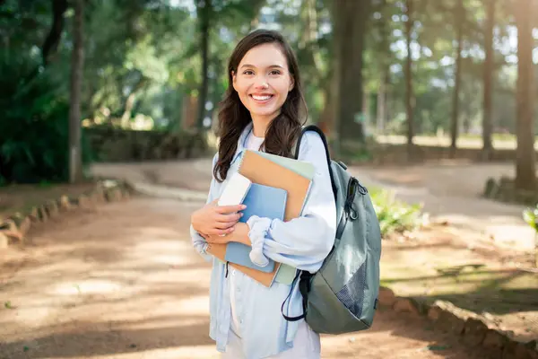 Beaming university european student lady holding educational materials and a smart device, poised in a sunlit park with a serene ambiance, symbolizing a blend of technology and learning