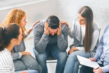 Alkolizm sorunu olan depresyonlu bir adam rehabilitasyon merkezinde oturuyor, grup onu destekliyor, boş yer var.