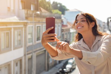 Güzel, genç bir kadın akıllı telefonuyla fotoğraf çekiyor. Balkonda dikilirken mutlu bir milenyum kadını şehir manzarasını yakalıyor, seyahatlerin ve yaz tatillerinin tadını çıkarıyor.