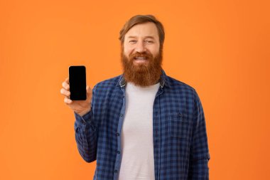 Harika bir uygulama. Pozitif kızıl sakallı adam akıllı telefonunun boş ekranını gösteriyor, mobil uygulama reklamı için model öneriyor, stüdyo turuncu arka planında boş bir cihaz dokunmatik ekranı ile poz veriyor.