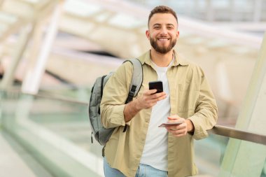 Havaalanında beklerken akıllı telefon kullanan yakışıklı bir gencin portresi, Terminalde sırt çantasıyla beklerken cep telefonuyla internete göz gezdiren mutlu yüzlü adam,