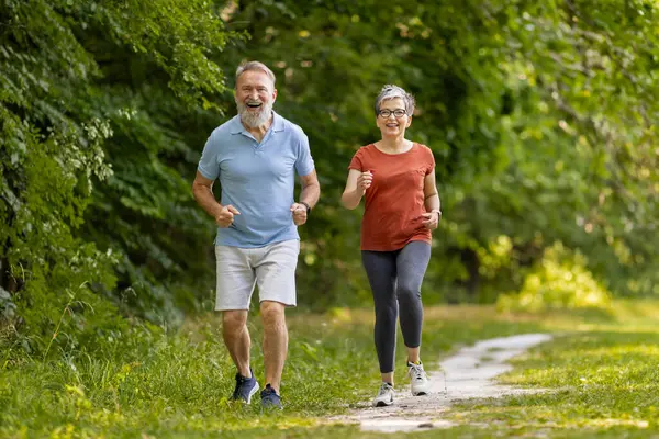 Joyful senior couple jogging together along park path, happy elderly man and woman in activewear running outdoors, enjoying healthy lifestyle and training outside, full length with copy space