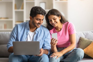 Genç Hintli çift gülümseyip akıllı telefona bakarken dizüstü bilgisayarda çalışan adam evdeki koltukta rahatça oturuyor.