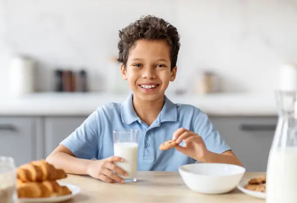 Happy black boy drinking milk and eating cookies in kitchen, holding glass, smiling. Cheerful kid keeping healthy diet nutrition, getting calcium, vitamins, probiotics for growth from dairy products