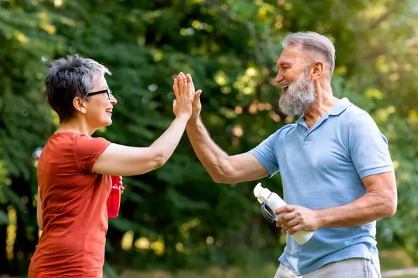 Energetic senior man and woman giving high-five to each other outdoors, happy cheerful elderly people celebrating fitness goals in sunny green park, holding water bottles in hand, side view