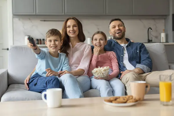 ハッピーヨーロッパの家族は 子供たちが家で一緒にテレビを見たり 若い両親と2人の子供を愛し リモートコントローラーを保持しているポップコーンとソファーでリラックス — ストック写真