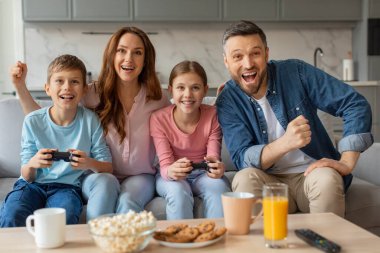 Evde video oyunu oynayan iki çocuklu neşeli genç aile, mutlu ergen kardeşler ve aileleri galibiyeti kutluyor, rahat oturma odalarında birlikte eğleniyorlar, kaynaşıyorlar ve gülüyorlar.