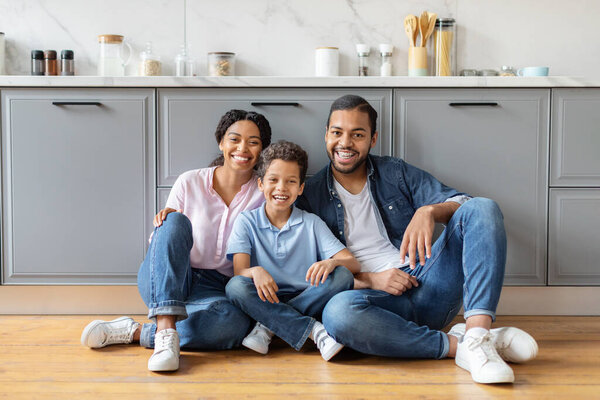 Portrait of loving african american family posing at kitchen. Happy beautiful mother, father and preteen boy son sitting on floor, embracing and smiling at camera. Childhood, love, affection