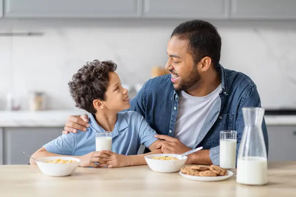 快乐的非洲裔美国父亲和未成年的儿子一起吃早餐 喝牛奶 吃谷物和做饭 爸爸抱着他的孩子 一边在厨房里吃饭一边聊天 — 图库照片