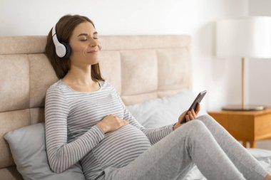 Rahat ve memnun bir şekilde, hamile bir kadın müzikten zevk alır ve yatakta dinlenirken akıllı telefonunu kullanır.