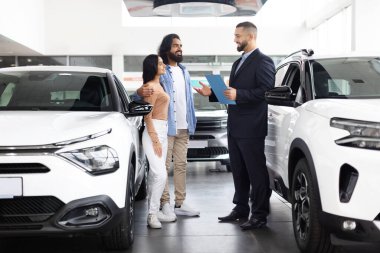 Hintli çift, yeni bir araç alımını göz önünde bulundurarak modern bir araba galerisinde bir araba satıcısıyla sohbet ediyor.