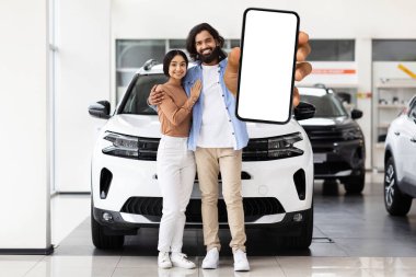 Heyecanlı Hintli çift, bir galeride beyaz ekranlı akıllı telefonu sunuyor. Dijital araba satın alma veya haber paylaşımı kavramı.