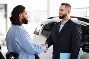 Takım elbiseli profesyonel bir satıcı ve Hintli bir müşteri el sıkışıyor. İyi aydınlatılmış modern bir araba galerisinde anlaşma yapıyor.
