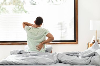 Afrika kökenli Amerikalı bir adam yatakta yatarken sırtını uzatıyor, uyandıktan sonra acı çekiyor, basit bir yatak odası düzeninde.