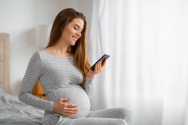 Lachende Zwangere Vrouw Surfen Haar Telefoon Zitten Een Gezellige Slaapkamer — Stockfoto
