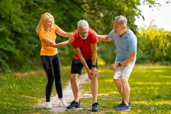 Concerned senior couple helping their male friend recover from injury during outdoor training, elderly man suffering muscle cramp or knee trauma after jogging in green park, free space