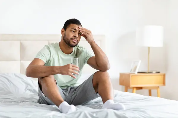 非裔美国人坐在床上 手里拿着一杯水 在舒适的家庭环境中显得疲惫或不舒服 — 图库照片