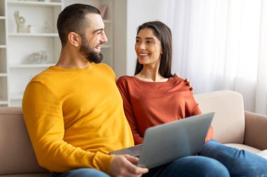 Neşeli bir adam ve kadın kanepede oturmuş laptoplarını paylaşıyor, birbirlerine eşlik ediyor ve eğleniyorlar.