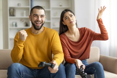 Neşeli bir adam ve şaşkın bir kadın birlikte video oyunları oynuyorlar, bir koltukta oturuyorlar, muhtemelen komik ve dostça bir şekilde yarışıyorlar.