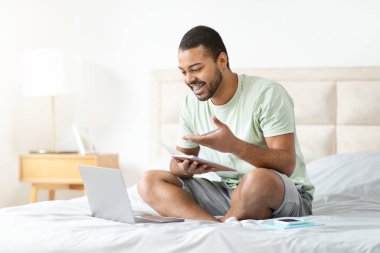 Afro-Amerikalı bir adam beyaz bir yatak örtüsüne dizüstü bilgisayarı ve not defteriyle oturuyor, evde çalışıyor gibi görünüyor.