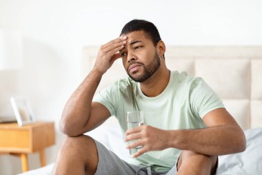 Afrika kökenli Amerikalı bir adam, eli alnında, elinde bir bardak su tutarak yatakta oturuyor. Sade bir ortamda baş ağrısı ya da endişeyi ifade ediyor.