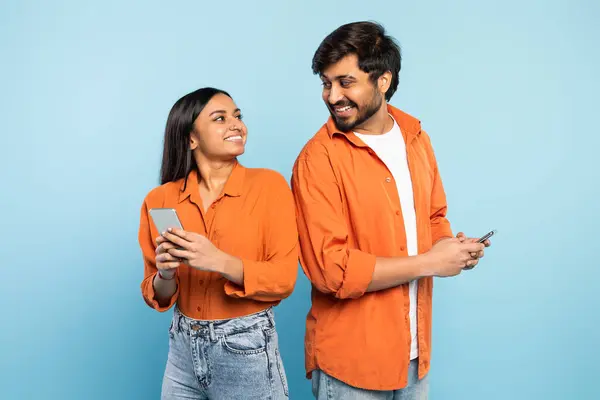 興奮したヒンドゥーのカップルは 背中に立ち スマートフォンを保持し 使用しながらお互いに微笑み ソーシャルネットワークのフィードをスクロールし スタジオで青い背景でオンラインチャット — ストック写真