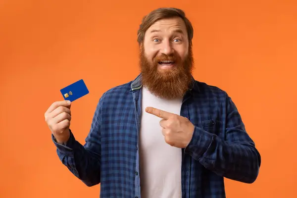 Excited Redhaired Bearded Guy Showing Credit Card And Pointing Finger, Advertising Bank Service Offer, Posing Over Orange Background In Studio. Concept Of Payment And Finances