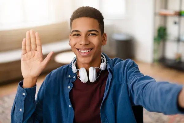 Happy black teenage guy with headphones around his neck taking selfie or having video call, waving hello to the camera with bright, friendly smile in home setting