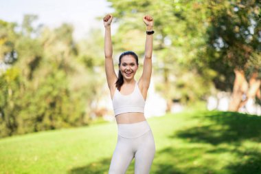 Coşkulu bir beyaz kadın kazanan kollarını kaldırarak spor salonunun başarısını kutlayan, beyaz bir spor kıyafeti ve saat takan, canlı bir park arka planına karşı, canlı bir park arkaplanına karşı.