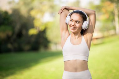 Neşeli, mutlu beyaz bir kadın beyaz kulaklıklarıyla müzik dinlerken kolunu uzatıyor. Arka planda yemyeşil bir parkta gülümsüyor.