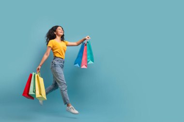 Renkli alışveriş torbalarıyla enerjik bir kadın canlı bir bakışla mavi bir arka planda zıplıyor.