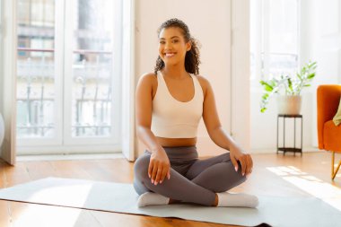 Spor yapan genç bir kadın, pilates yapıyor. Nilüfer pozisyonunda oturuyor. Modern ev spor salonunda yoga seansına hazır.