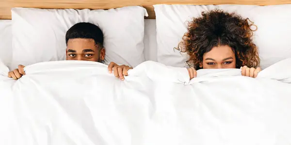 Funny millennial african-american couple hiding under white blanket in bed, top view
