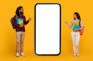 Genç, modaya uygun giyinen erkek ve kadın sarı arka planda boş bir akıllı telefon görüntüsüne işaret ediyor.