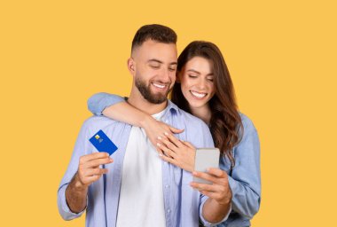 Kredi kartı ve akıllı telefon tutan neşeli çift online alışverişi sembolize ediyor.