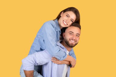 Bir kadının bir erkeğin sırtına bindiğini gösteren neşeli bir görüntü. İkisi de sarı arka plana gülümsüyor. Eğlenceyi ve oyunculuğu simgeliyor.