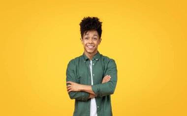 Amiable young black man with arms crossed, flashing a charming smile in front of a sunny yellow background clipart