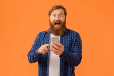Kızıl saçlı iş adamı, akıllı telefonda modern uygulama kullanırken şaşkınlığını dile getiriyor. Turuncu stüdyo arka planında heyecanlı bir şekilde kameraya bakıyor. Mobil teknoloji ve eğlence