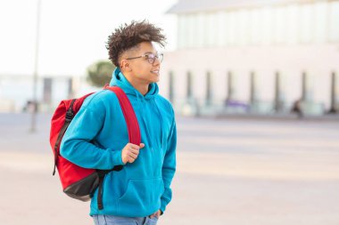 Confident young brazilian guy student walking in an urban setting with a red backpack and looking into the distance clipart